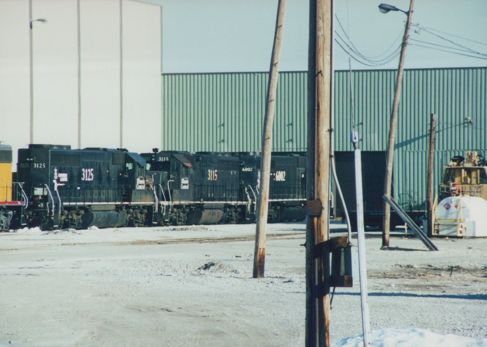 Three black diesel locomotives with white frame stripes stand in a row in front of a large green corrugated metal building, with snow on the ground and wooden utility poles in the foreground.