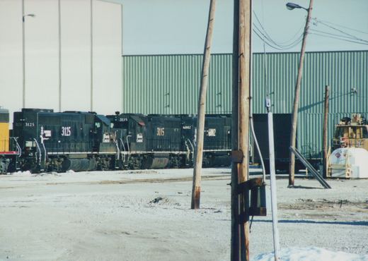 Three black diesel locomotives with white frame stripes stand in a row in front of a large green corrugated metal building, with snow on the ground and wooden utility poles in the foreground.