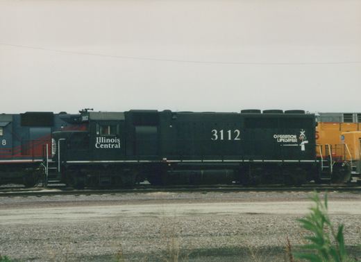Side view of a black Illinois Central diesel locomotive numbered 3112 with an Operation Lifesaver emblem, coupled between a blue and red locomotive and a yellow locomotive in a gravel rail yard under an overcast sky.