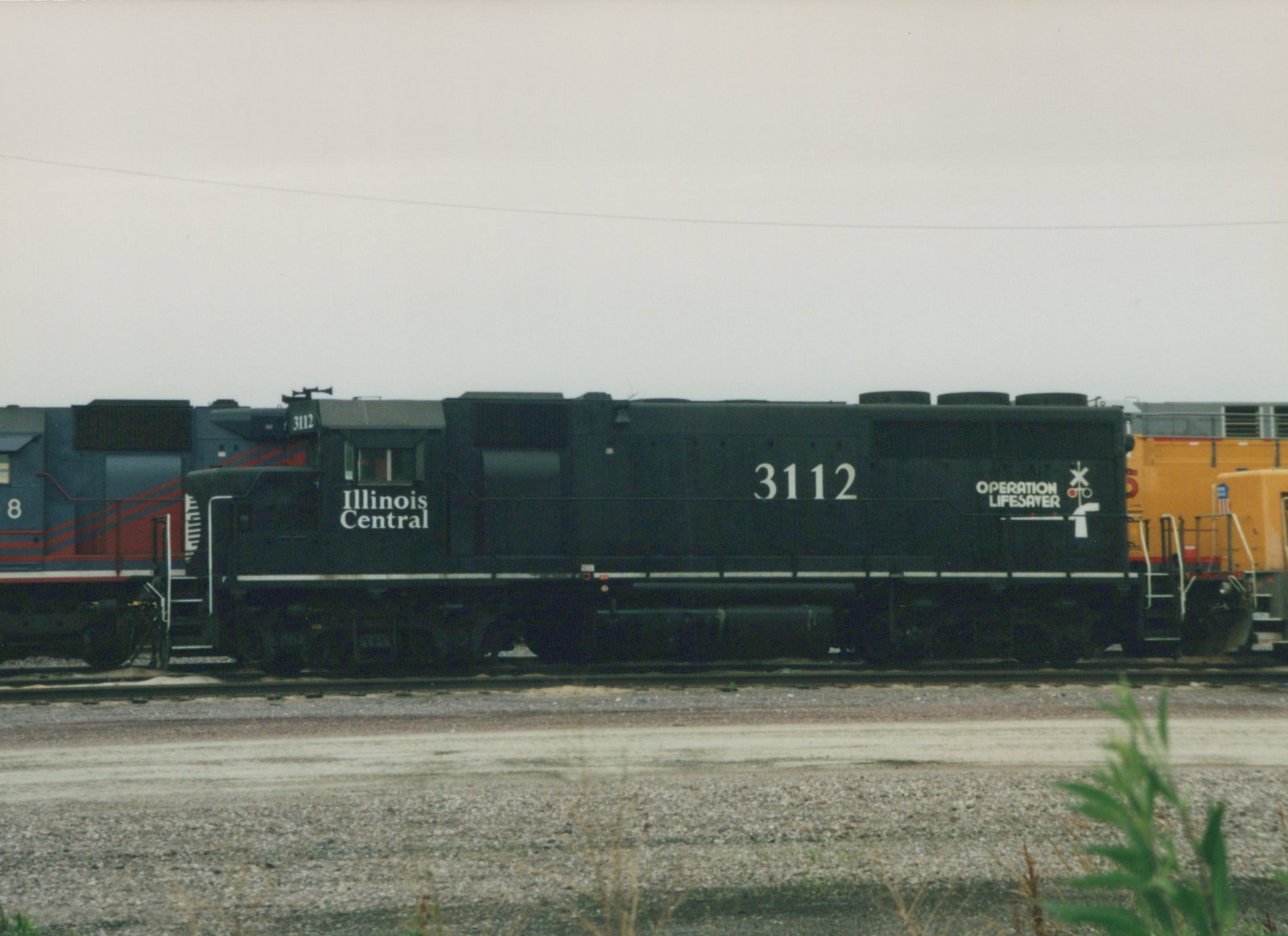 Side view of a black Illinois Central diesel locomotive numbered 3112 with an Operation Lifesaver emblem, coupled between a blue and red locomotive and a yellow locomotive in a gravel rail yard under an overcast sky.