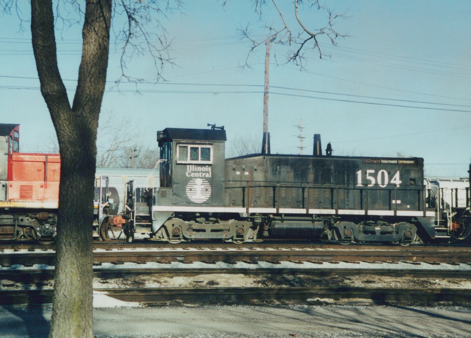 Side view of a black Illinois Central switcher numbered 1504 on a yard track, with a white herald on the cab, a bare tree in the foreground, a red-orange locomotive at the left and a white covered hopper at the right.