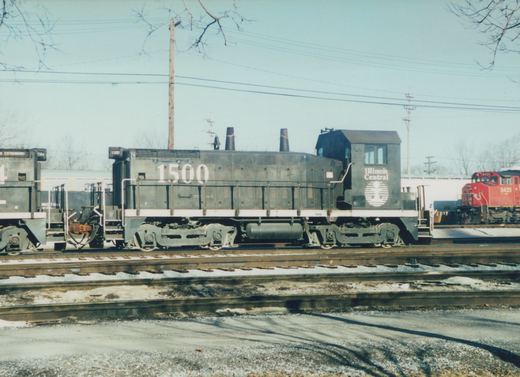 A black Illinois Central end-cab diesel switcher numbered 1500 stands in a snowy rail yard beside a second black switcher, with a red locomotive numbered 5435 in the distance.