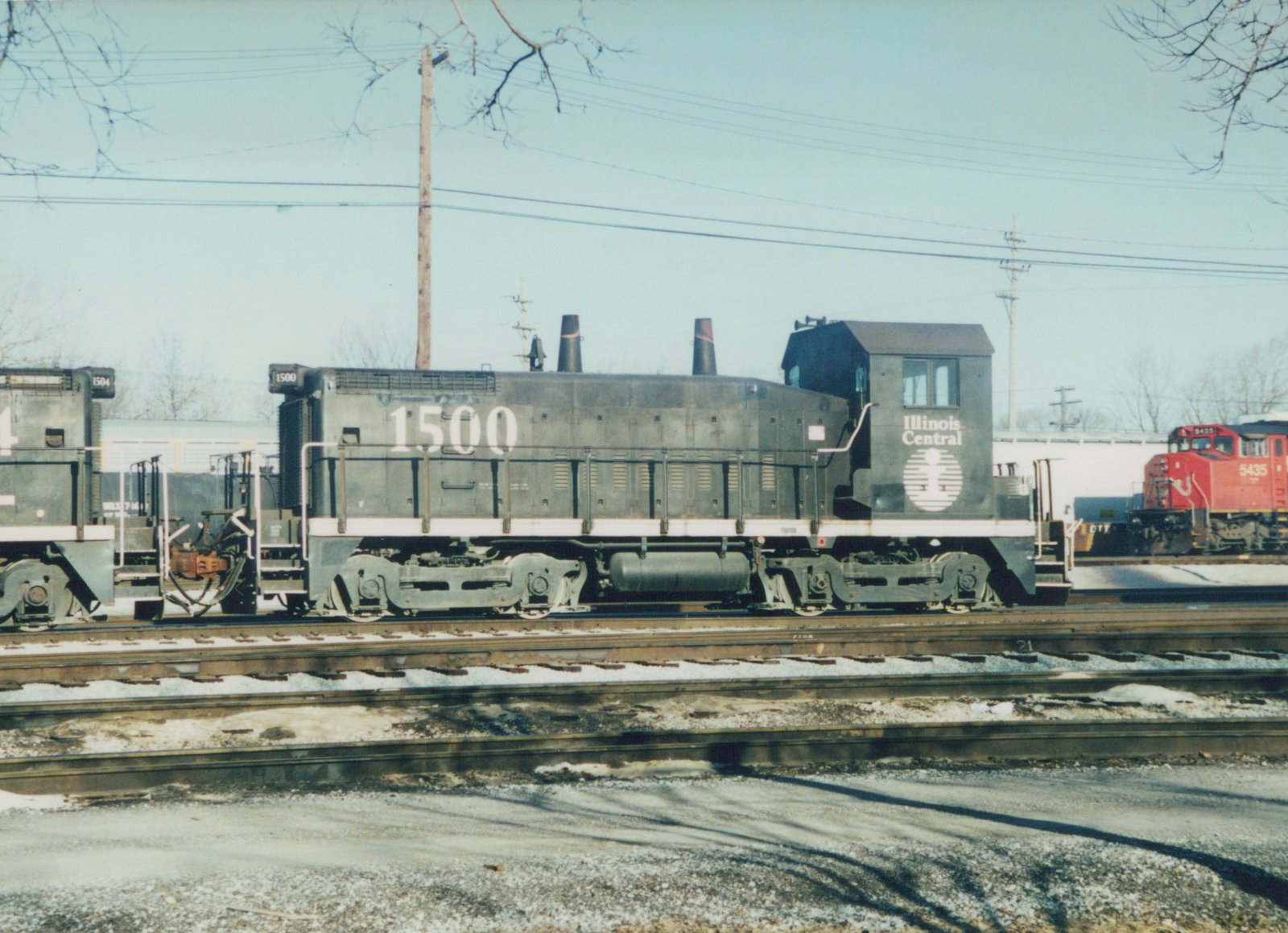 A black Illinois Central end-cab diesel switcher numbered 1500 stands in a snowy rail yard beside a second black switcher, with a red locomotive numbered 5435 in the distance.