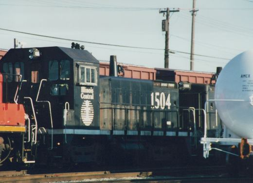 Side view of a black diesel switcher lettered Illinois Central and numbered 1504, with a second black switcher behind it, a white tank car at right and the red-orange nose of another locomotive at left.