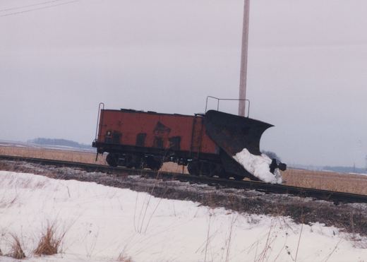 An orange railroad snowplow with a large black wedge blade packed with snow stands on a single track, with a snowy embankment in the foreground and flat fields and an overcast sky behind it.