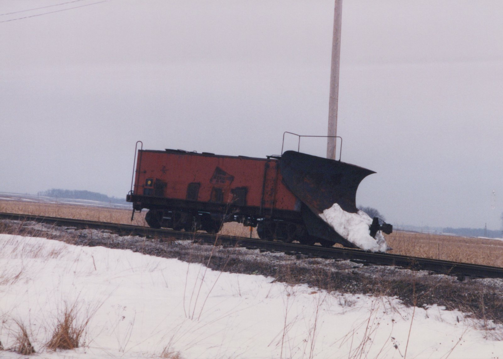 An orange railroad snowplow with a large black wedge blade packed with snow stands on a single track, with a snowy embankment in the foreground and flat fields and an overcast sky behind it.