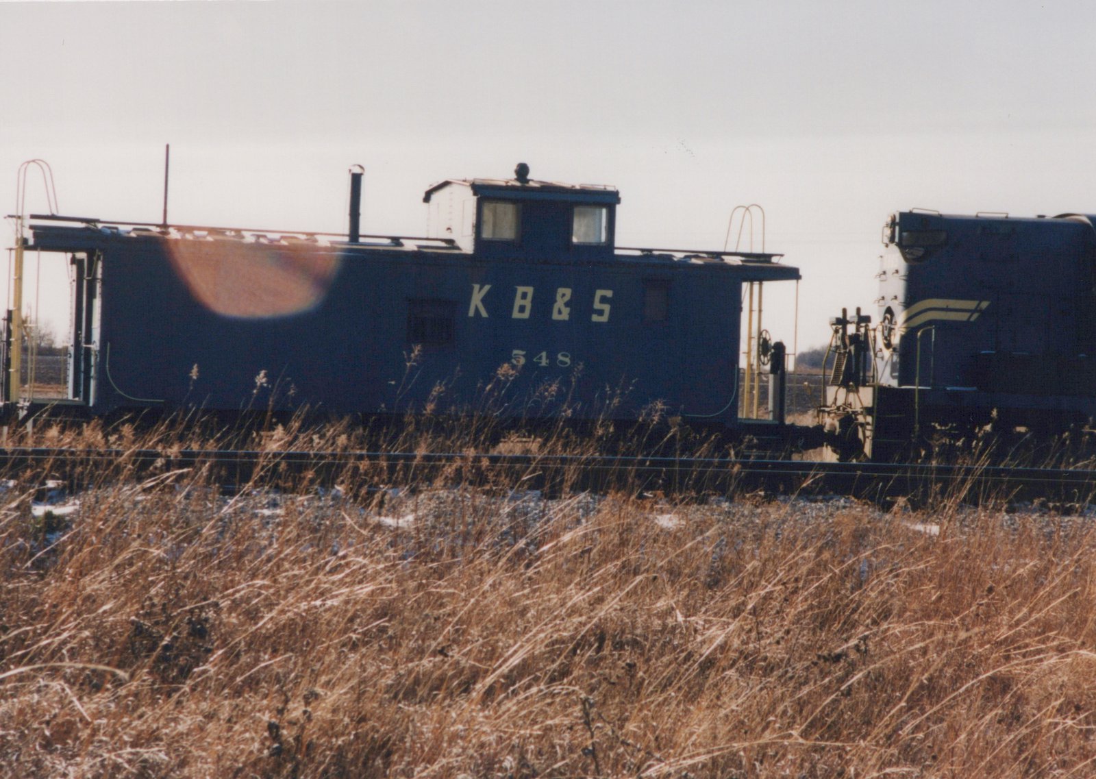 A dark blue caboose with a cupola, lettered K B & S and numbered 548, stands on a track beside the end of a blue diesel locomotive with cream stripes, with tall dry grass and a light dusting of snow in the foreground.
