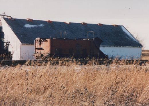 A faded red railroad snow plow with a wedge blade sits on a track in front of a long white shingle-roofed building, with the end of a dark locomotive at the left and tall dry grass in front.