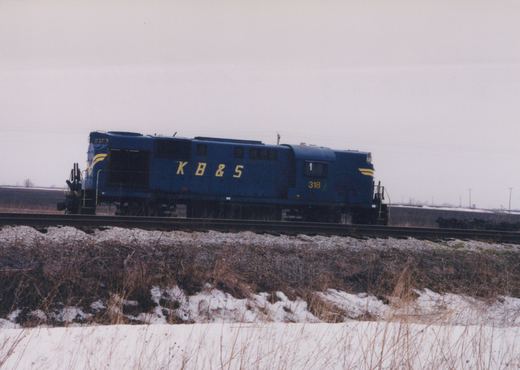 A blue diesel locomotive lettered K B & S in yellow, number 318 on the cab, stands on a single track with snow and dry grass in the foreground under a gray sky.
