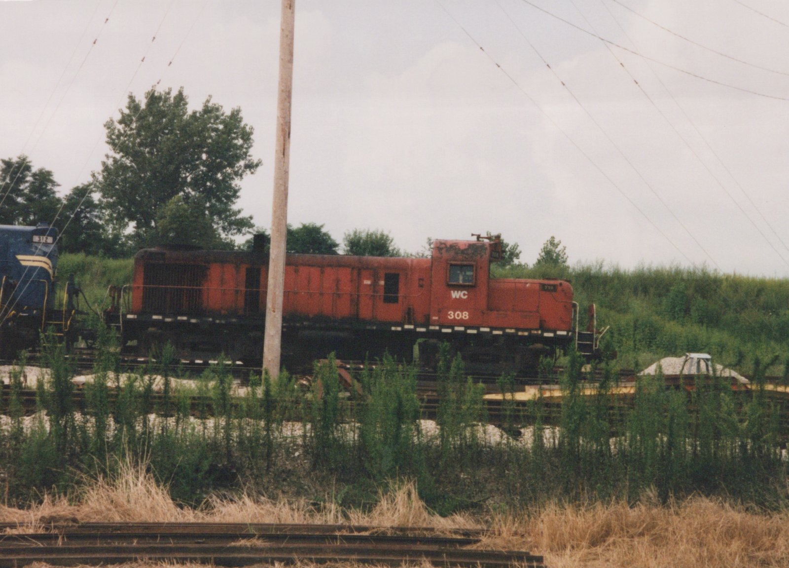 Side view of a faded, rust-streaked red diesel switcher lettered WC 308 on a track behind tall weeds, with the end of a blue locomotive numbered 312 at left and a utility pole in front.