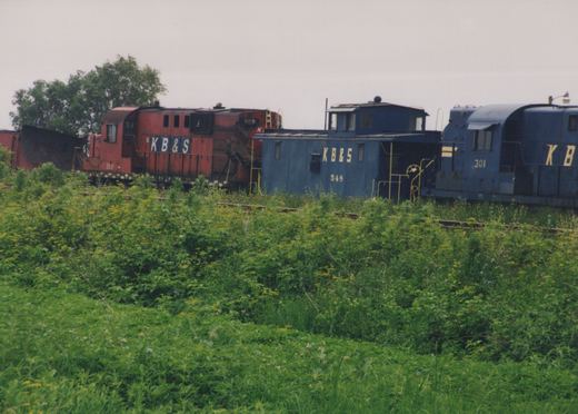 A red diesel locomotive with a snowplow, a blue caboose and a blue diesel locomotive, all lettered KB&S, on a weedy track behind a field of green brush under a gray sky.