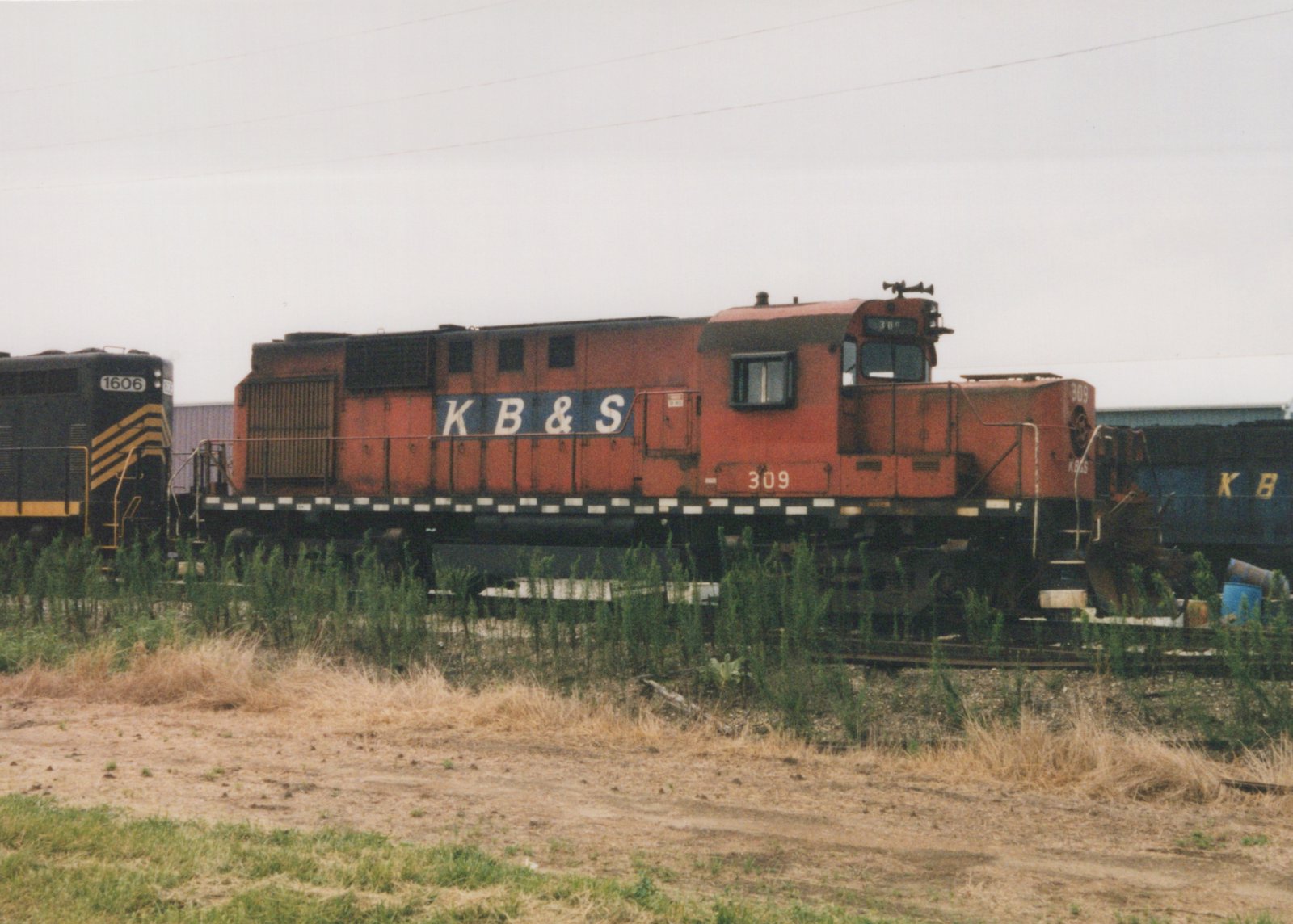 A faded red-orange diesel locomotive lettered K B & S and numbered 309 is parked on a weedy track under an overcast sky, between a black and yellow locomotive numbered 1606 and a blue locomotive.