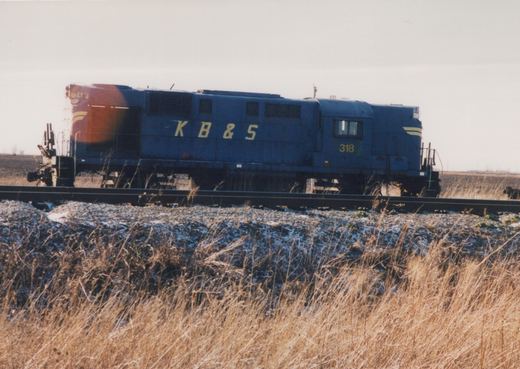 Side view of a dark blue diesel road-switcher locomotive lettered K B & S, with number 318 on the cab, on a single track above a snow-dusted ballast bank and dry grass, with flat fields and a hazy sky behind.