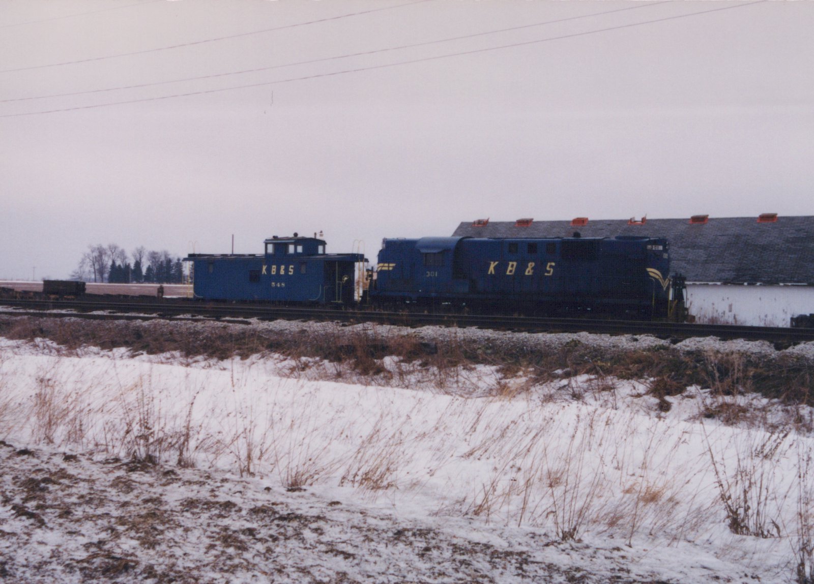 Side view of a dark blue diesel locomotive lettered KB&S and numbered 301 coupled to a blue caboose, on a track beside a snowy, grassy embankment under an overcast sky, with a long barn-like building behind them.