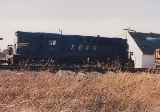 A dark blue diesel road switcher lettered K B & S and numbered 301 stands on a track behind a bank of tall dry grass, with a white gabled building and the end of a rust-red freight car on the right.