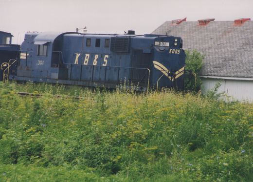 A dark blue diesel locomotive lettered KB&S and numbered 301, with cream chevrons on its hood end, sits on a weedy track in front of a white wooden building with a shingled roof.