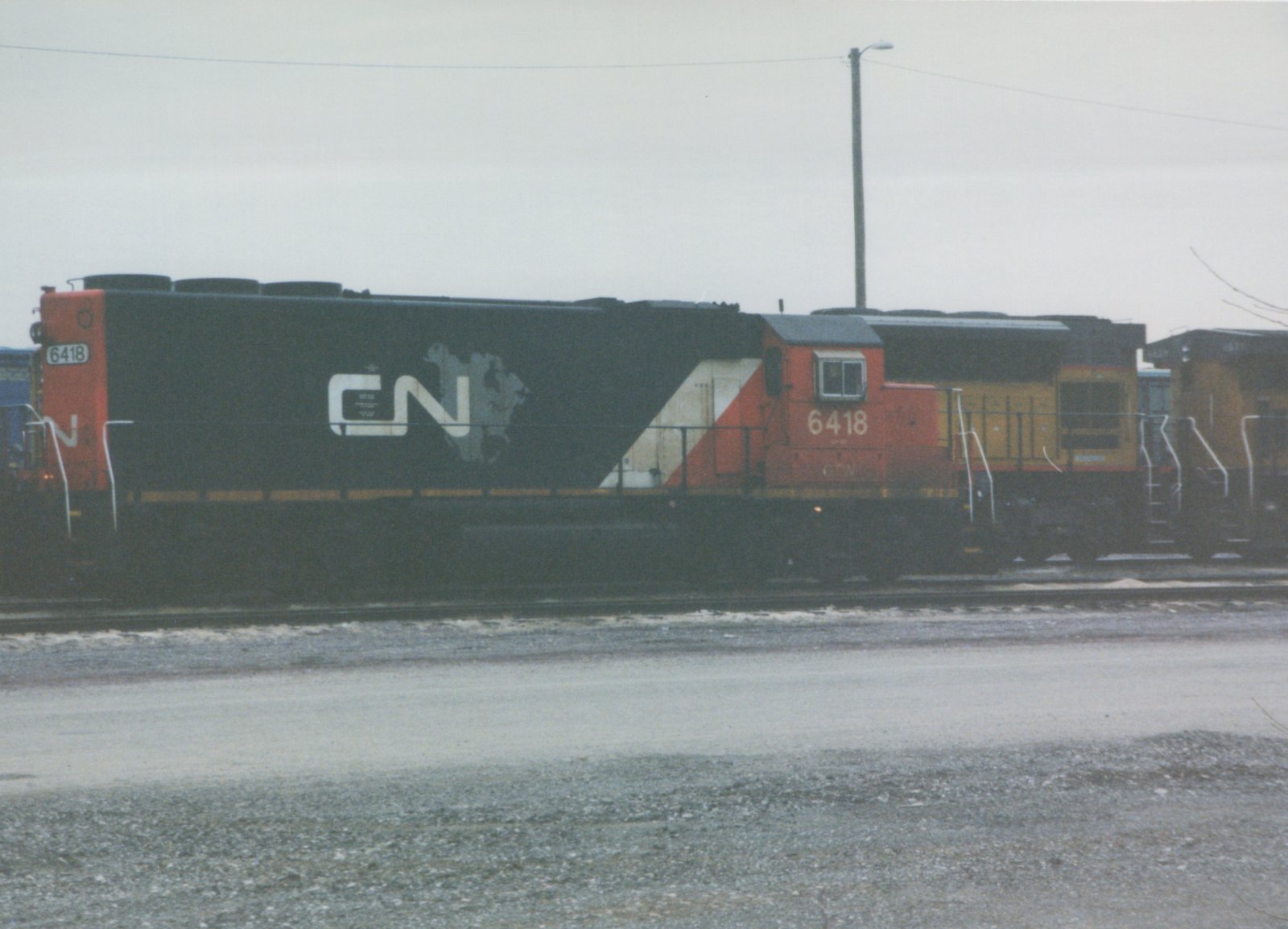 Side view of a black and red CN diesel locomotive numbered 6418, coupled to yellow locomotives on a yard track under a grey sky.