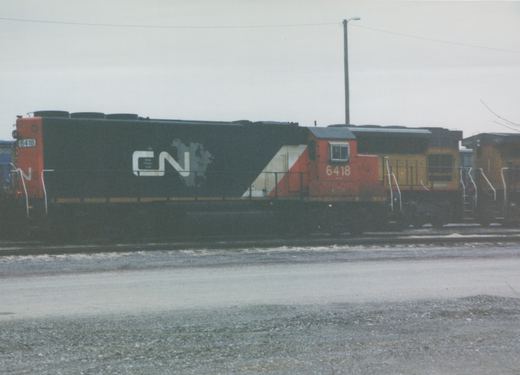 Side view of a black and red CN diesel locomotive numbered 6418, coupled to yellow locomotives on a yard track under a grey sky.