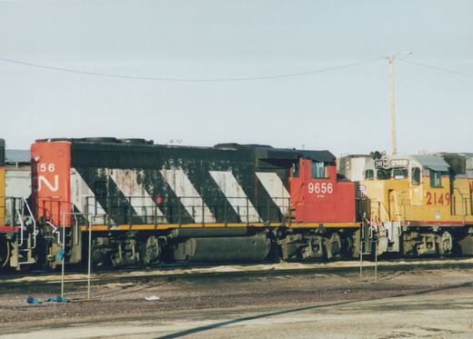 Side view of a red, black and white striped diesel locomotive numbered 9656, coupled near a yellow locomotive numbered 2149 on a yard track under a pale sky.