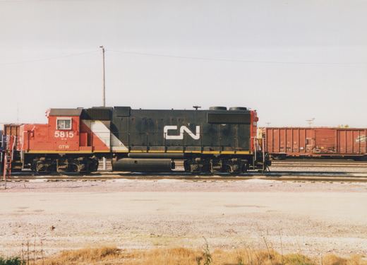 Side view of a red, white and black diesel locomotive numbered 5815 with a large white CN logo, standing on a yard track with a red Chicago and North Western boxcar behind it on the right.