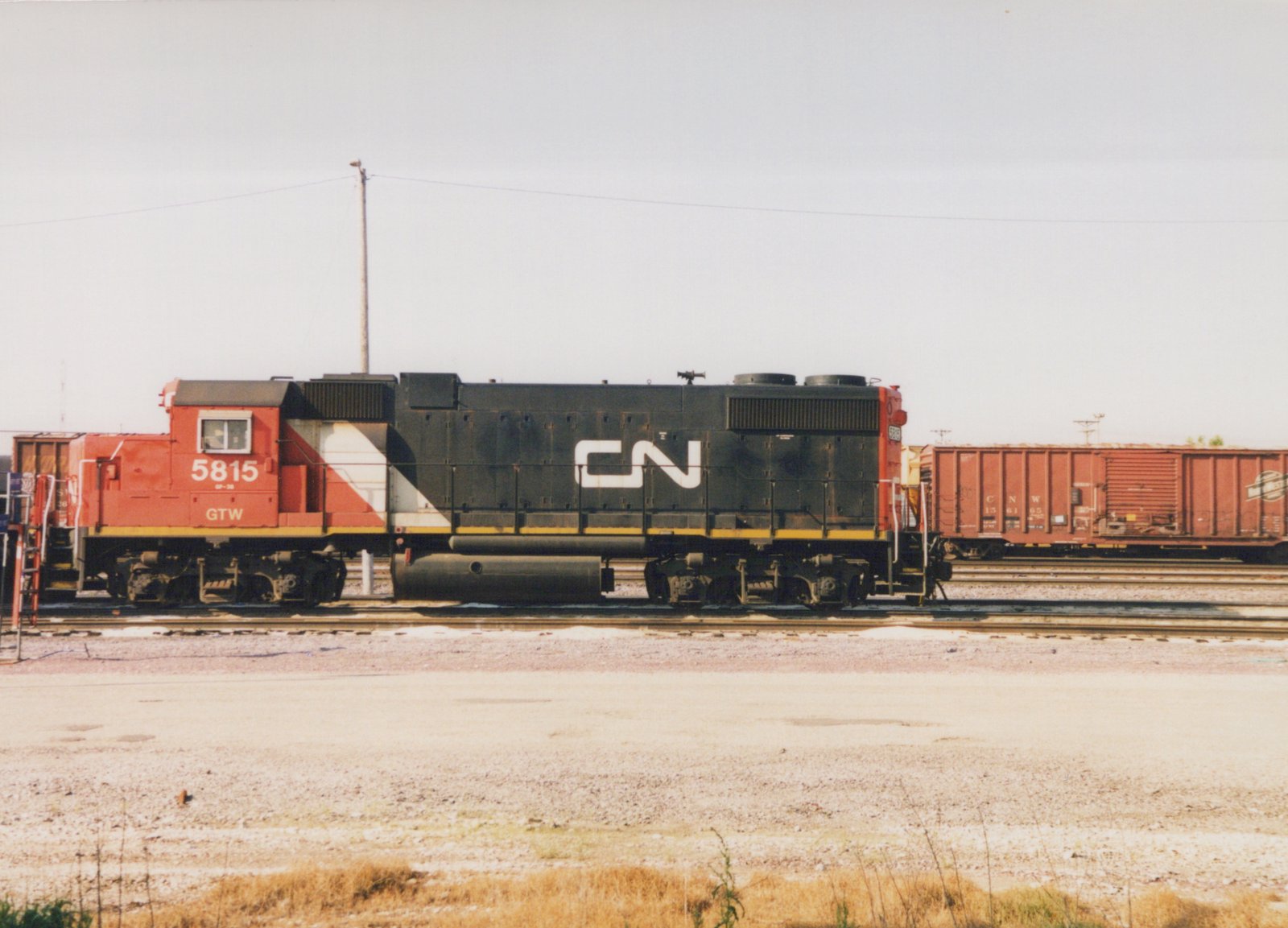 Side view of a red, white and black diesel locomotive numbered 5815 with a large white CN logo, standing on a yard track with a red Chicago and North Western boxcar behind it on the right.
