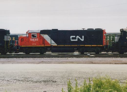 Side view of a diesel locomotive with a red cab and a black hood carrying a large white CN logo, parked between two black locomotives on a yard track under a gray sky.
