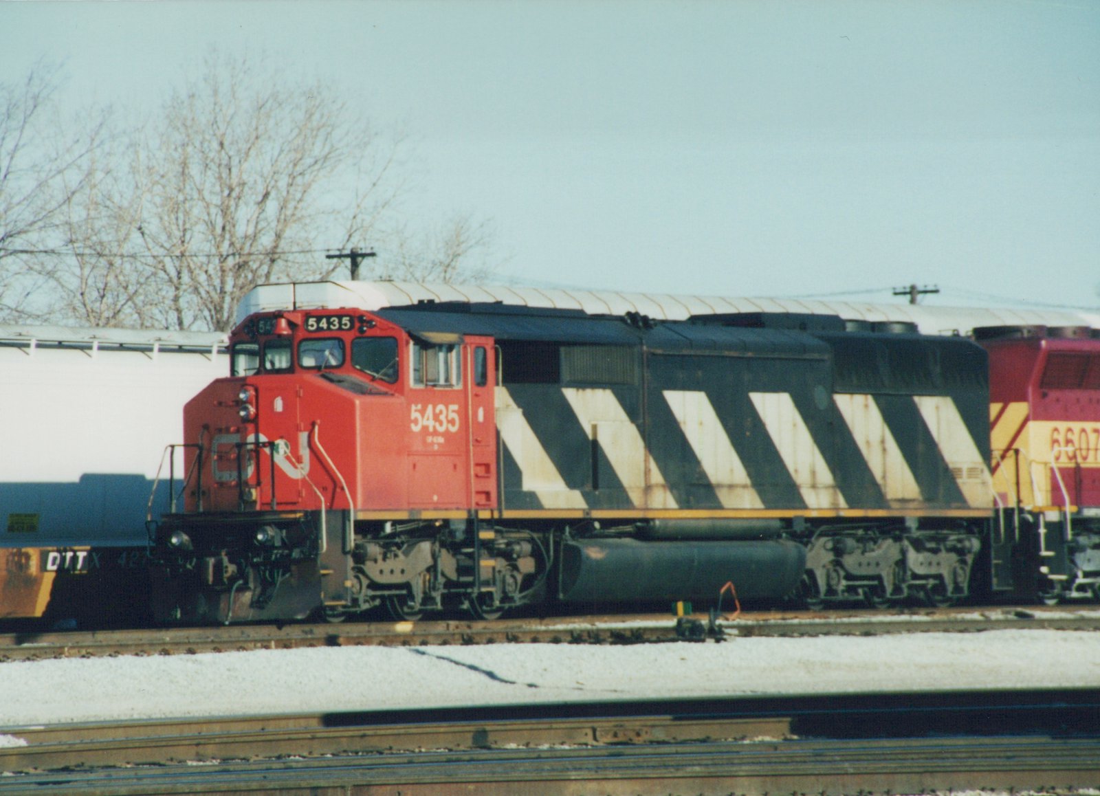 A red, black and white striped Canadian National diesel locomotive numbered 5435 on a yard track in snow, with a maroon and gold locomotive behind it and a white autorack beyond.