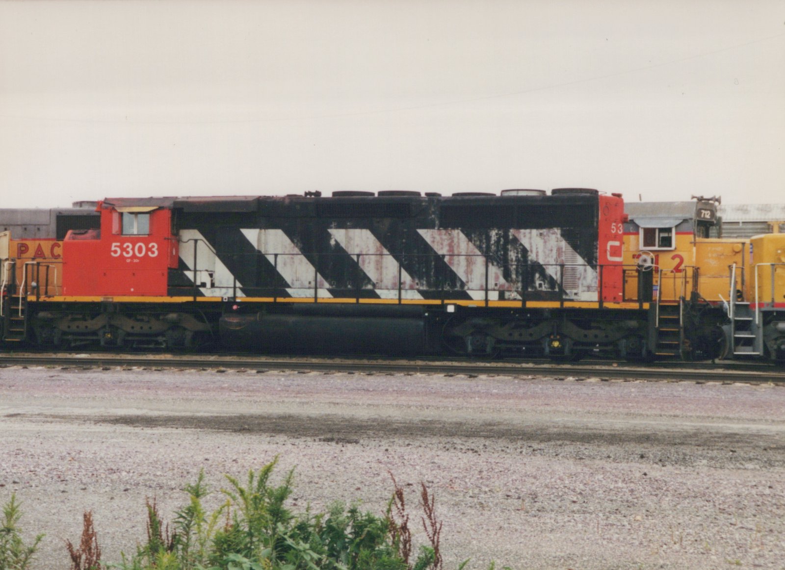 Side view of a weathered red, black and white diesel locomotive numbered 5303 on a yard track, coupled between yellow locomotives, under an overcast sky with weeds in the foreground.