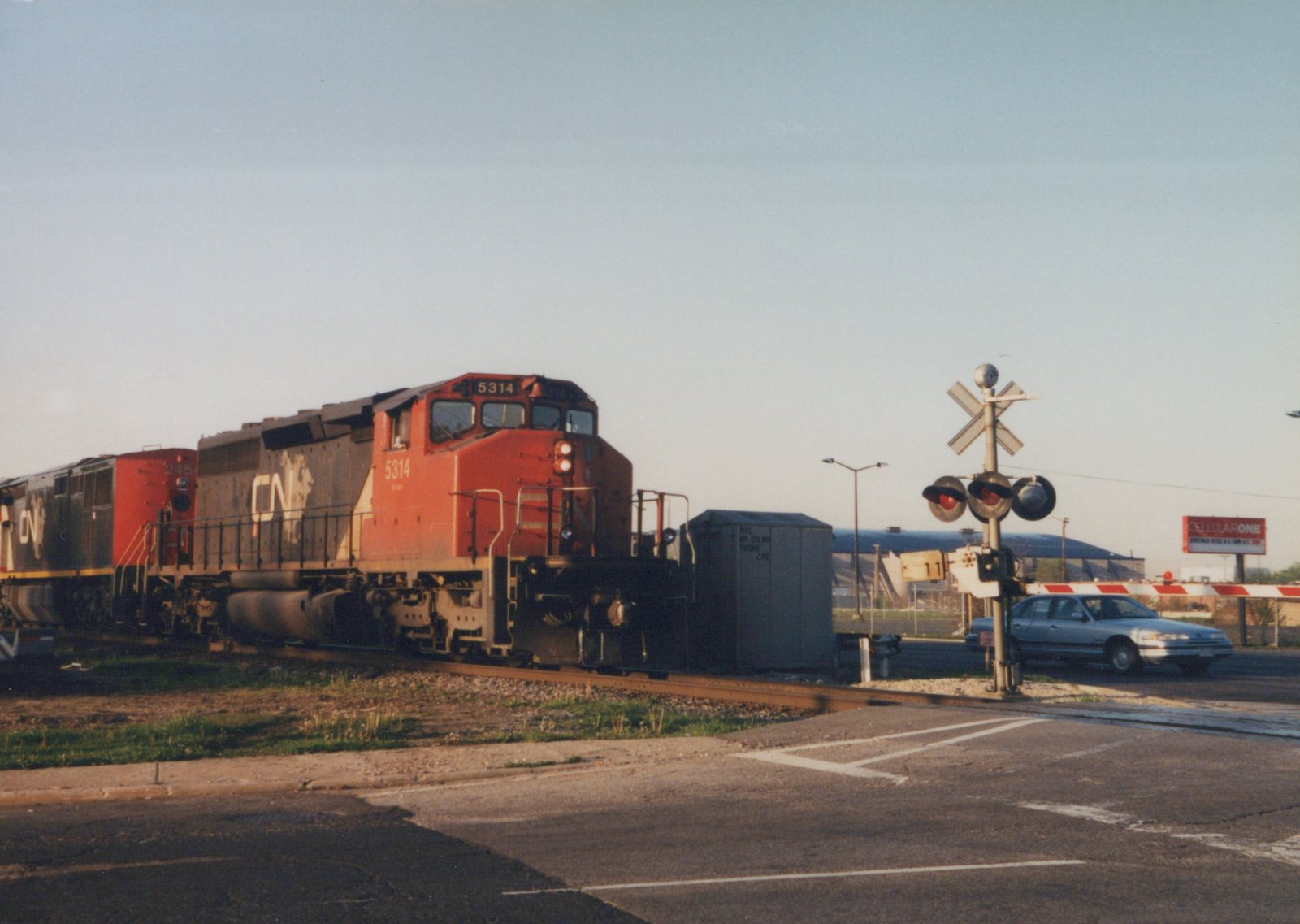 A red and black Canadian National diesel locomotive numbered 5314 leads two more locomotives past lowered crossing gates and flashing lights while a silver sedan waits at the crossing.