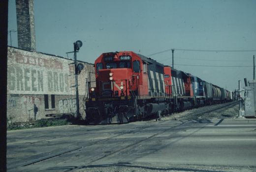 A red-orange and black diesel locomotive with a CN logo leads three more locomotives and a line of freight cars across a paved street, beside an old brick building with a faded GREEN BROS wall sign.