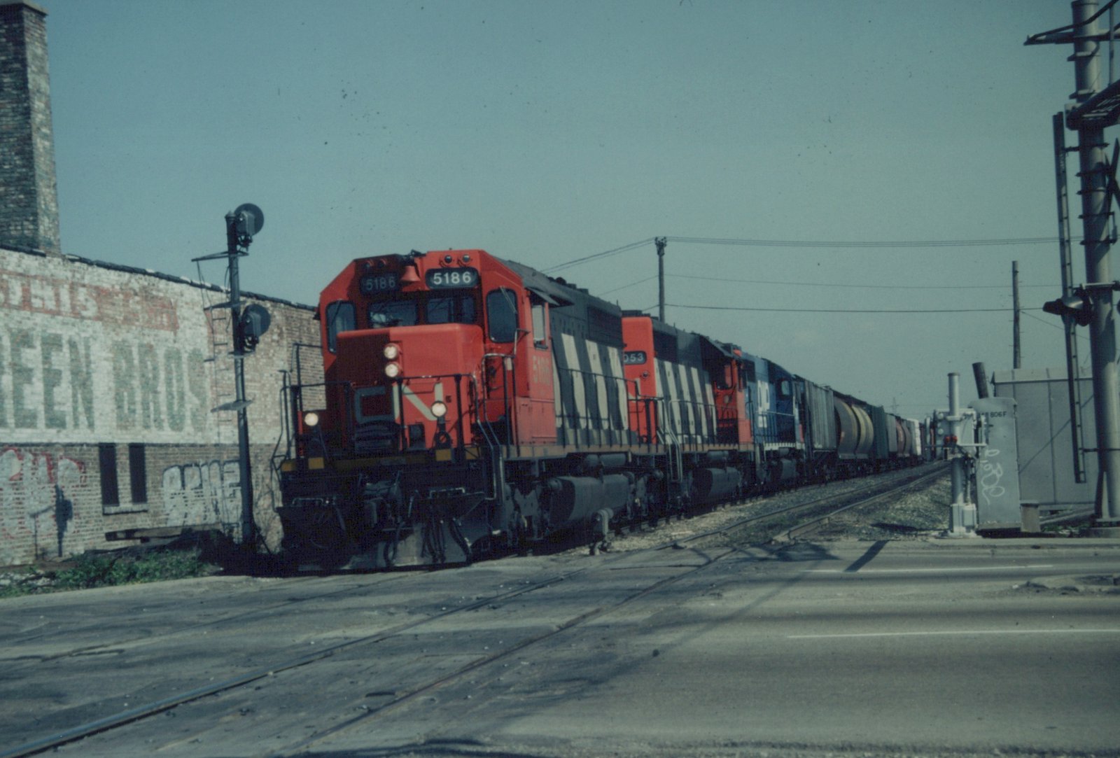 A red-orange and black striped diesel locomotive numbered 5186 leads three more locomotives and a long freight train across a paved street, beside an old brick building with a faded painted sign.