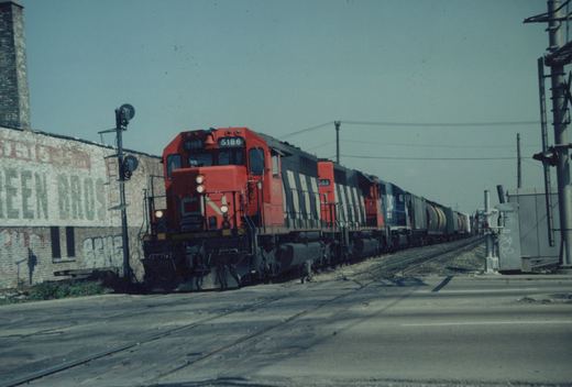A red-orange and black striped diesel locomotive numbered 5186 leads three more locomotives and a long freight train across a paved street, beside an old brick building with a faded painted sign.