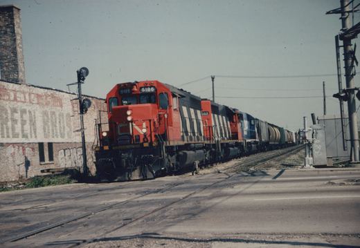 A red-orange and black Canadian National diesel locomotive with white diagonal stripes leads three more locomotives and a long freight train over a paved grade crossing beside an old brick building with a faded painted sign.