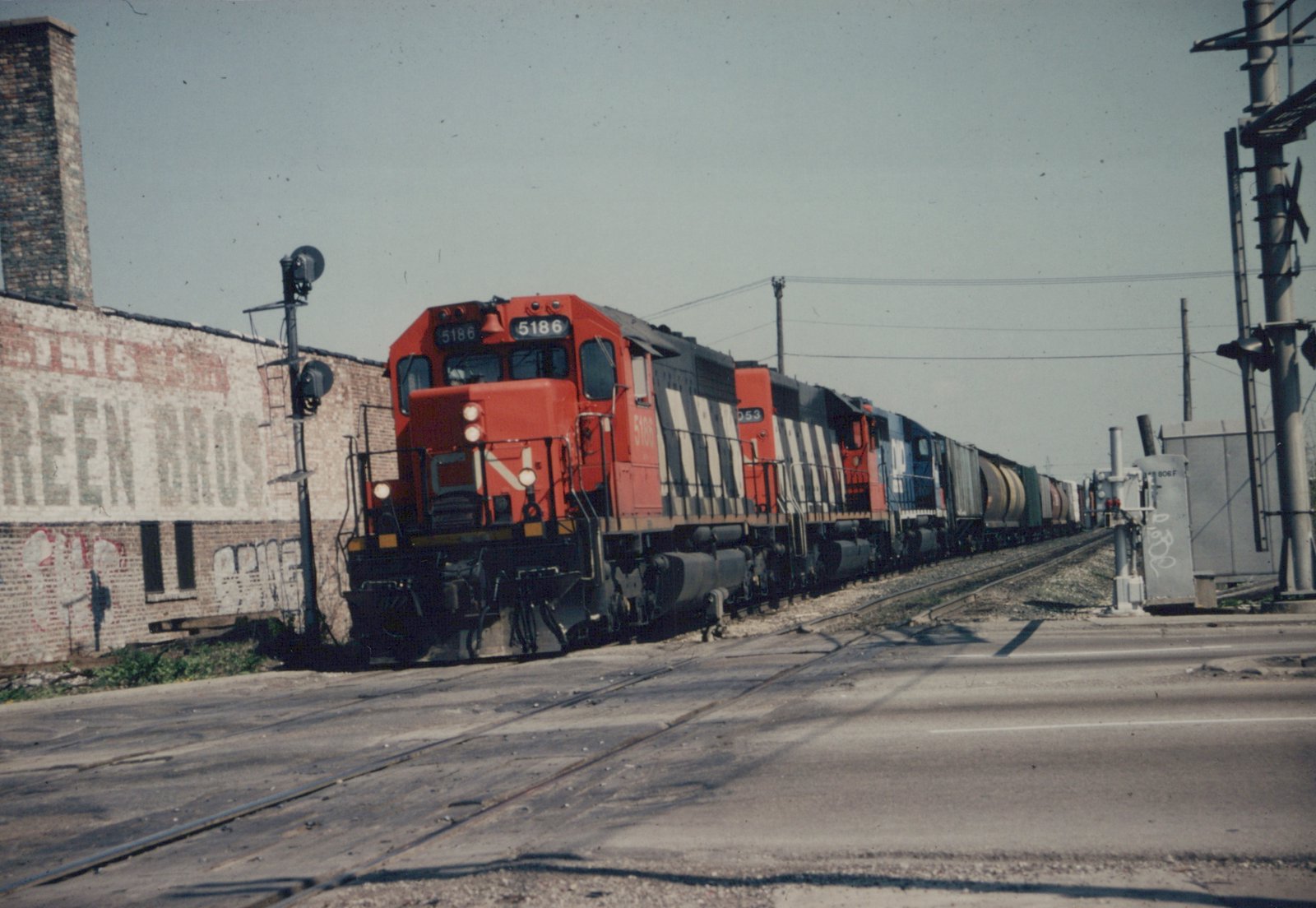 A red-orange and black Canadian National diesel locomotive with white diagonal stripes leads three more locomotives and a long freight train over a paved grade crossing beside an old brick building with a faded painted sign.
