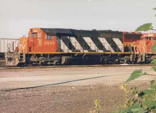 A red-orange and black diesel locomotive with white diagonal stripes on its hood, numbered 5049, stands on a yard track with two more locomotives behind it, seen across a gravel roadway with leafy weeds in the right foreground.