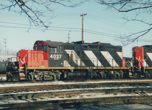 Side view of a black diesel locomotive with white diagonal stripes, a red cab and nose, numbered 4027, with a second similar locomotive behind it, on yard tracks under bare tree branches.