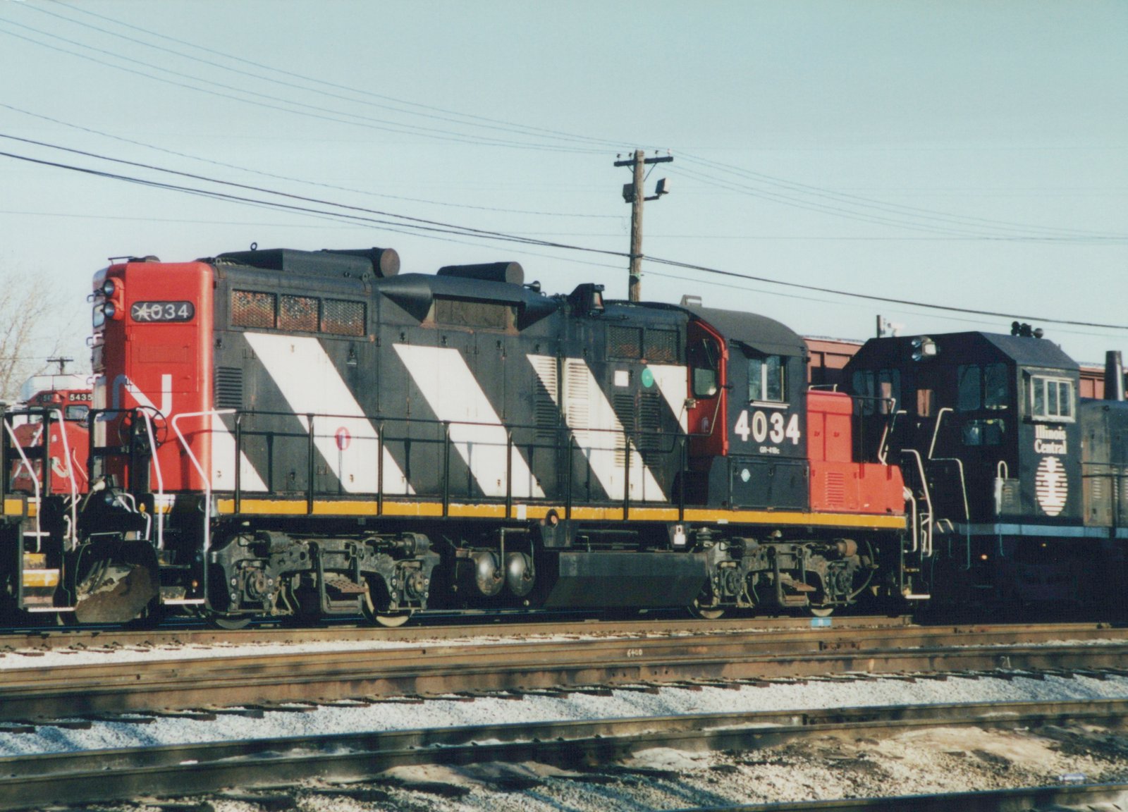 Side view of a black locomotive with white diagonal stripes and a red cab and nose, numbered 4034, on a yard track, with a black Illinois Central switcher to its right and a red locomotive behind at left.