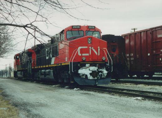 A red and black CN diesel locomotive numbered 2606, with snow packed on its pilot, stands on a track under bare tree branches beside a dark red freight car.