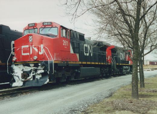 A red and black CN diesel locomotive with snow packed on its front pilot, followed by two more CN units, beside a gravel road under bare trees.