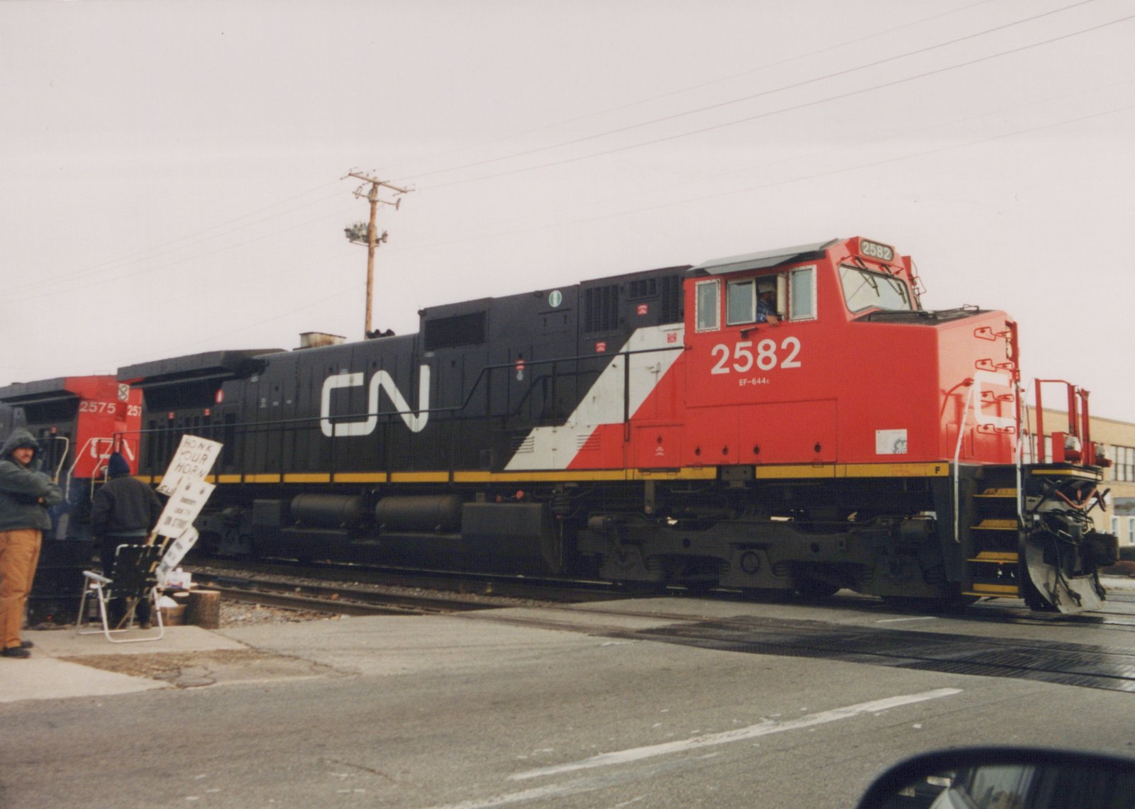 A red, white and black CN diesel locomotive numbered 2582, with a second CN unit numbered 2575 behind it, crosses a street at a grade crossing while two people with hand-lettered picket signs stand beside the track on an overcast day.