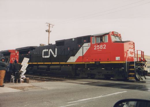 A red, white and black CN diesel locomotive numbered 2582, with a second CN unit numbered 2575 behind it, crosses a street at a grade crossing while two people with hand-lettered picket signs stand beside the track on an overcast day.