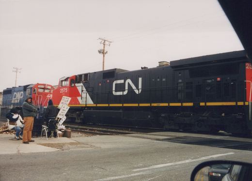 A black and red CN diesel locomotive numbered 2575 crosses a street, while three picketers with hand-lettered strike signs wait beside the tracks and a blue and red DWP locomotive stands behind them.