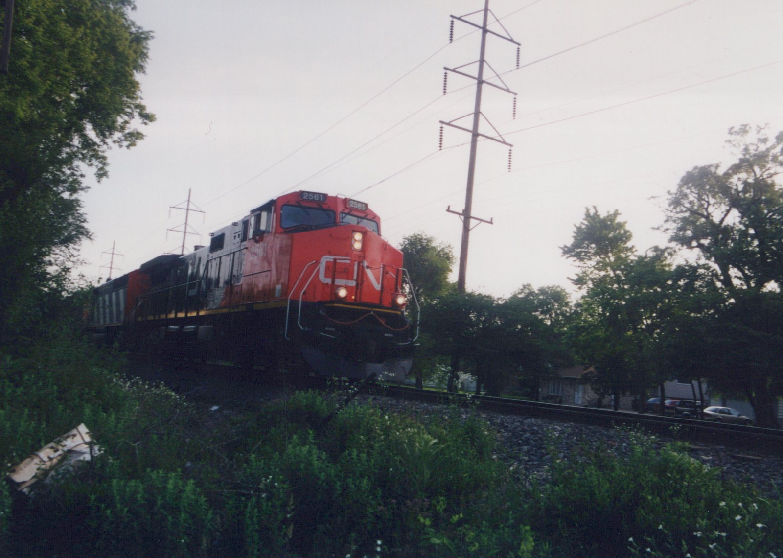 A red Canadian National diesel locomotive with a white CN logo on its nose and its headlights on approaches along a single track. A second locomotive follows, and a transmission-line pole rises above trees and weeds.