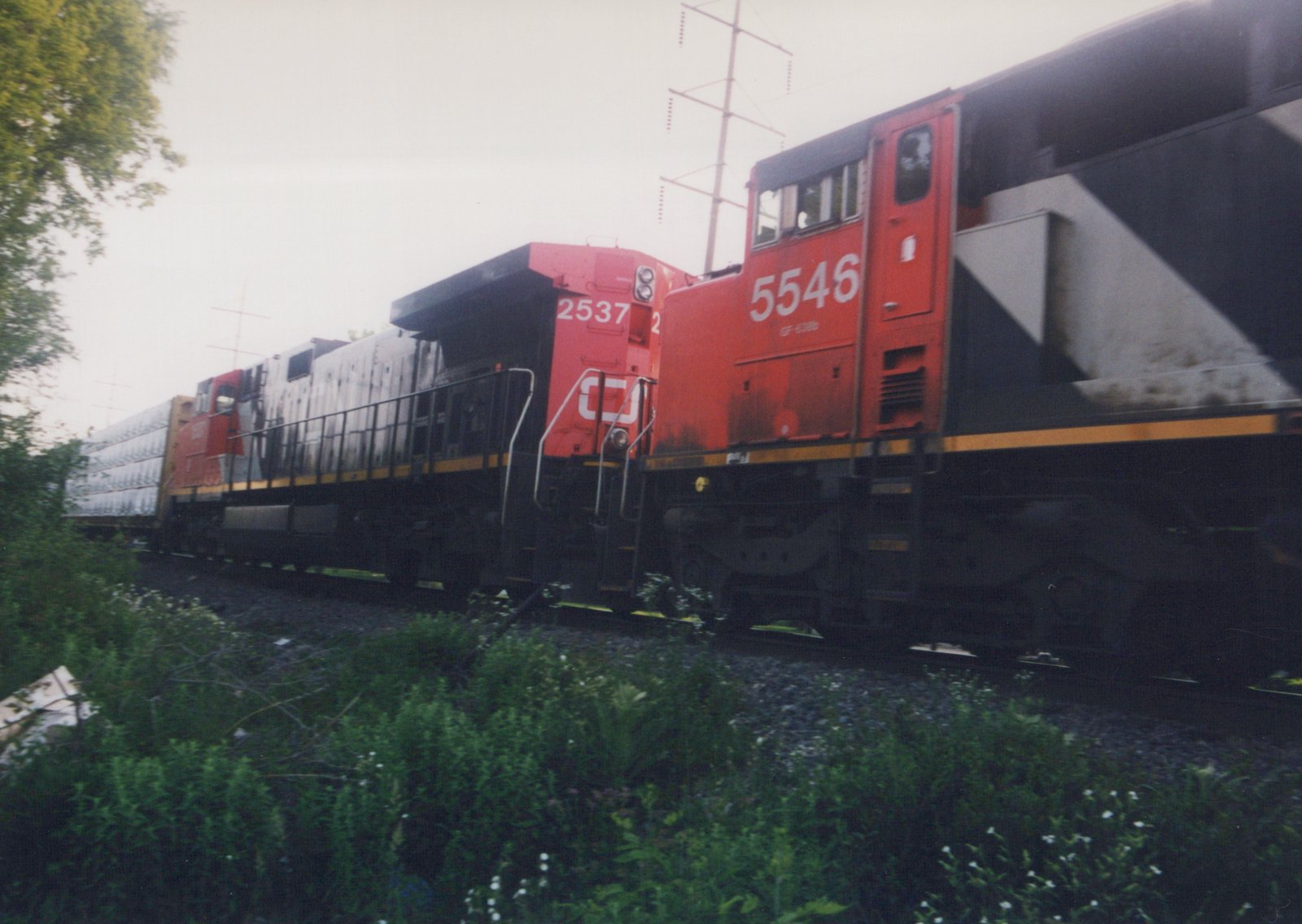 A moving freight train led by a red-orange and black locomotive numbered 5546, coupled to the rear of locomotive 2537 with the CN logo, on a weedy track under power line towers.