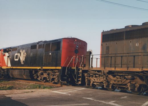 A black and red CN diesel locomotive numbered 2454, coupled long hood to long hood with a faded gray locomotive, crosses a paved road in low sunlight.