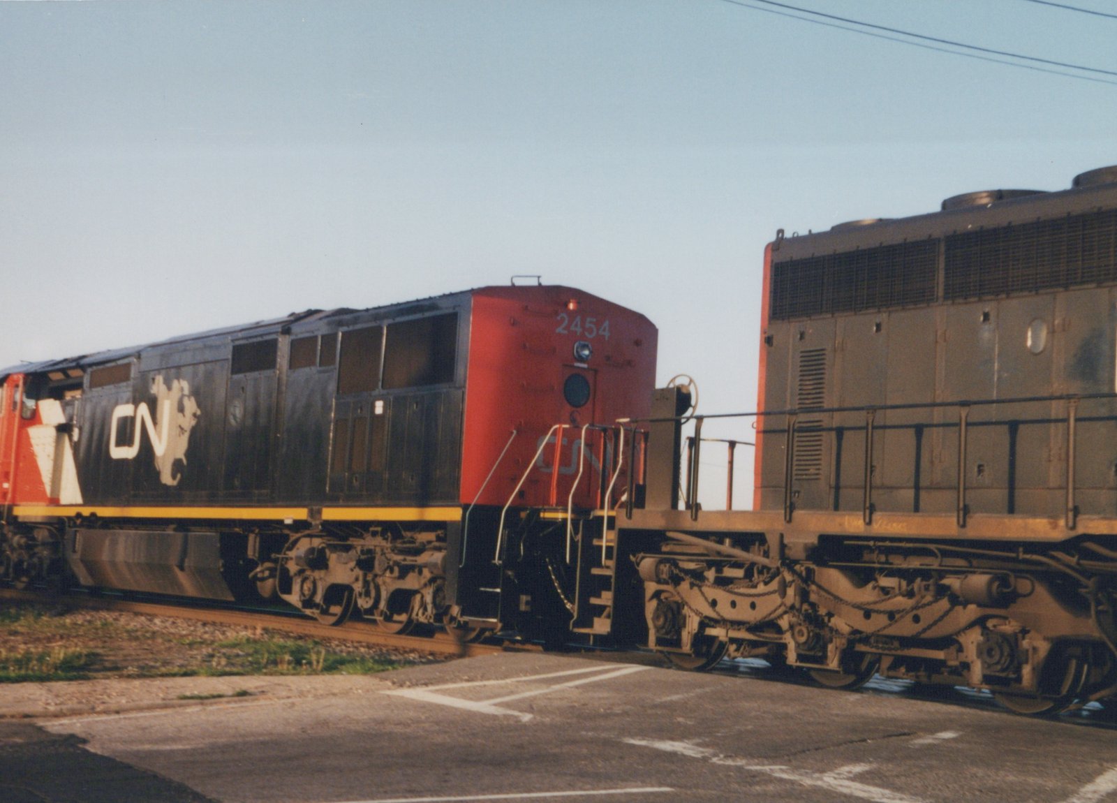 A black and red CN diesel locomotive numbered 2454, coupled long hood to long hood with a faded gray locomotive, crosses a paved road in low sunlight.