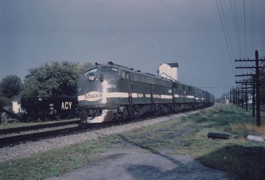 A dark green diesel locomotive with a white stripe and a CENTRAL herald on its nose leads a line of units and cars along a double-track line past a black ACY hopper car, with a white grain elevator and a pole line under a gray sky.