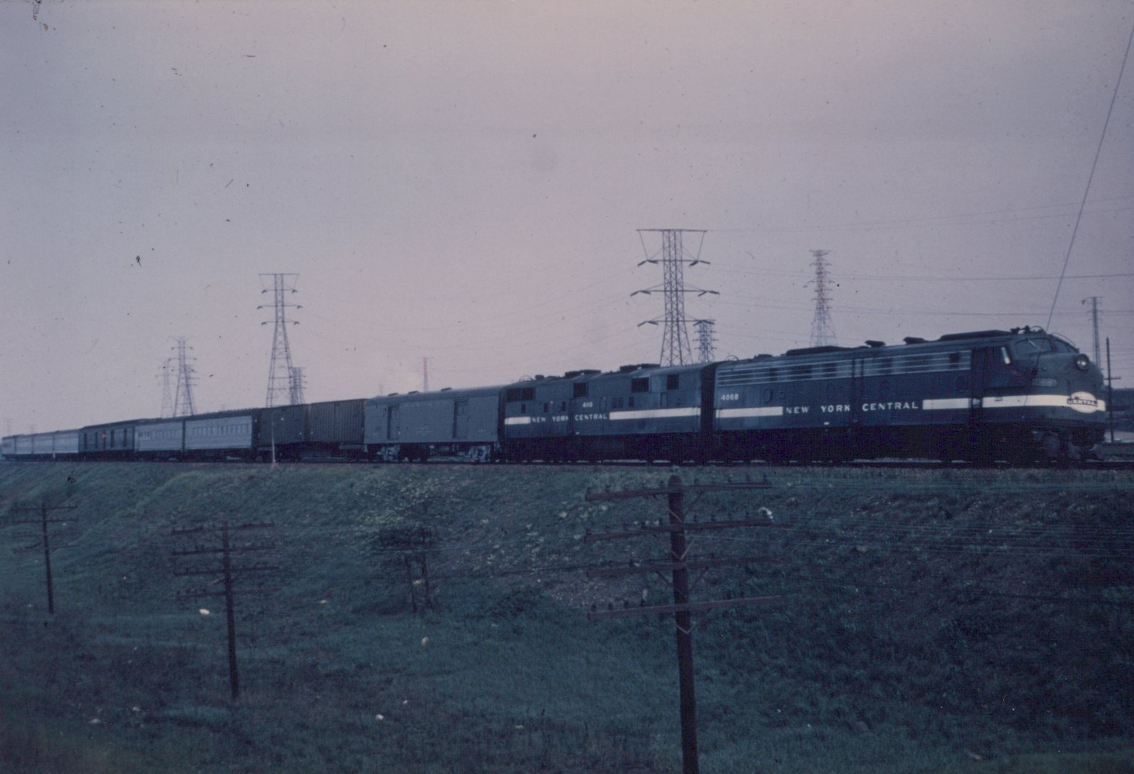 A dark gray New York Central streamlined diesel and a hood-type diesel pull a long train of baggage, express and passenger cars along a grassy embankment under transmission towers on an overcast day.