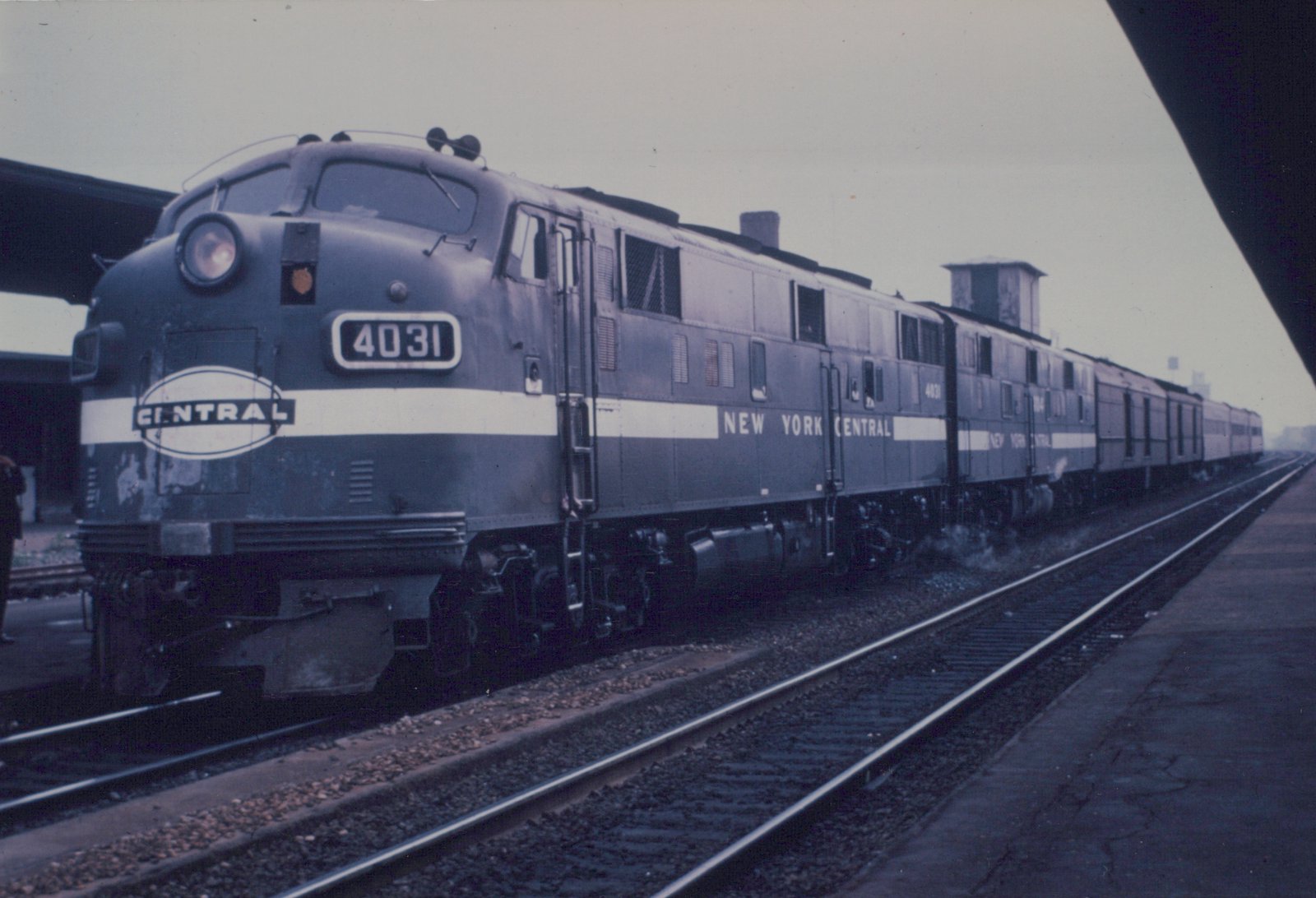 A dark gray New York Central streamlined diesel locomotive numbered 4031, with a white band and an oval CENTRAL herald, stands with a second unit and a train of passenger cars beside a station platform on an overcast day.