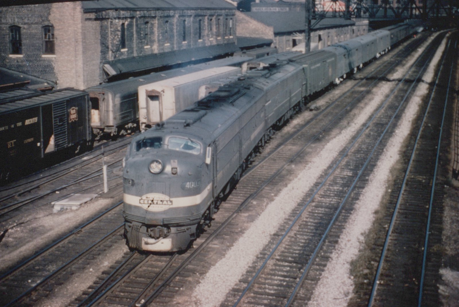A gray streamlined diesel locomotive with a CENTRAL herald on its nose and a booster unit behind it pulls a line of express cars along multiple tracks past brick freight houses.